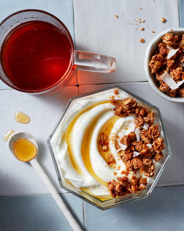 An octagonal glass bowl of yogurt and granola topped with sweet and spicy ginger syrup shown with a glass mug of syrup, a bowl of granola, and a mixing spoon. Sweet and Spicy Ginger Syrup