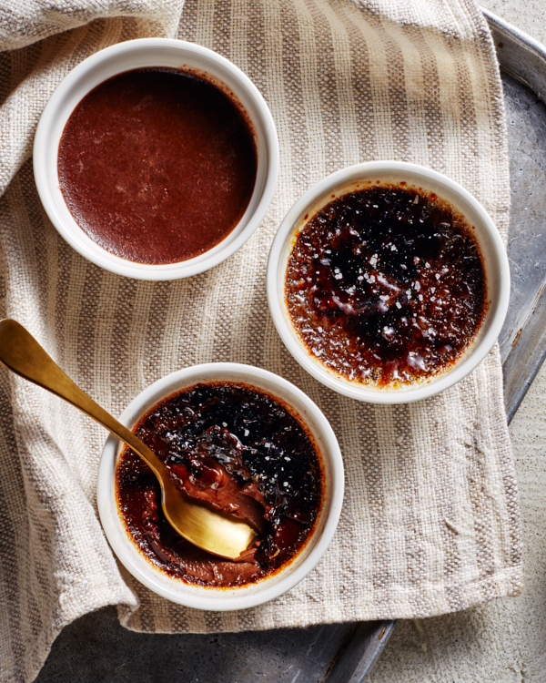 Three ramekins of salted dark chocolate crème brûlée, one partially eaten, one with no topping, shown on a tea towel on a baking tray with a gold spoon and a bowl of Golden Yellow sugar.