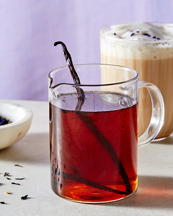 A glass pitcher of vanilla simple syrup with a vanilla bean pod, shown with a glass mug of London Fog Latte and a dish of loose Earl Grey tea