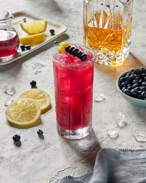 Blueberry Sortilege Whisky Fizz A highball glass of blueberry whisky fizz on ice garnished with a skewer of blueberries, shown with a decanter of whisky, a pitcher of simple syrup, lemon wedges and wheels, and a bowl of blueberries.
