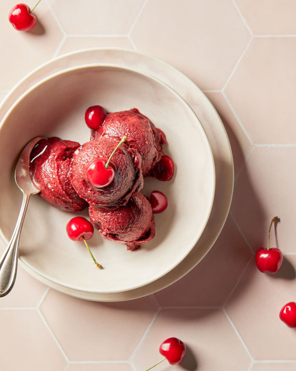 4-Ingredient Cherry Sorbet Four scoops of cherry sorbet garnished with cherries, shown on a counter of pale pink hexagonal tiles.